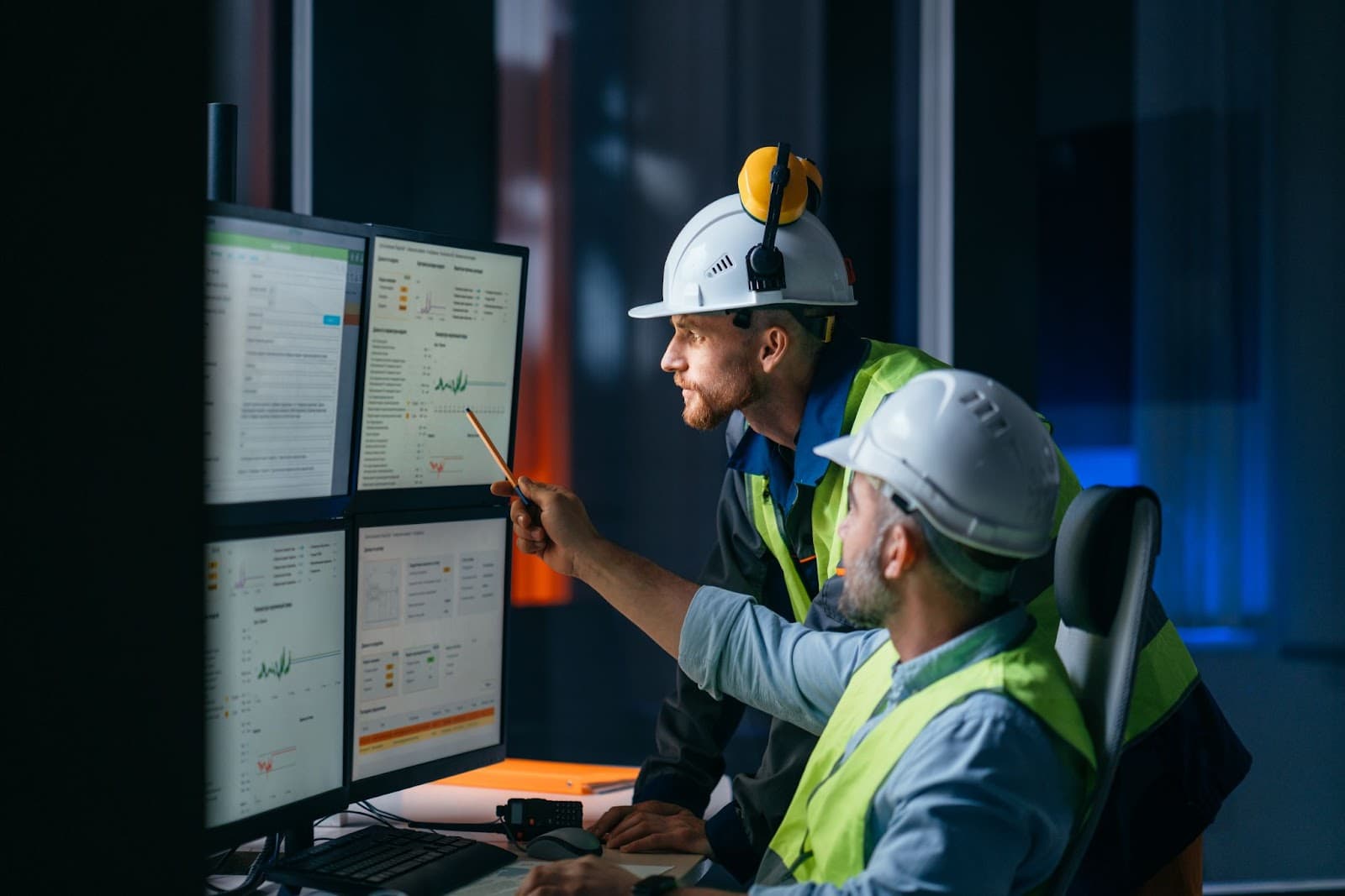 Two men wearing hard hats looking at four computer screens.